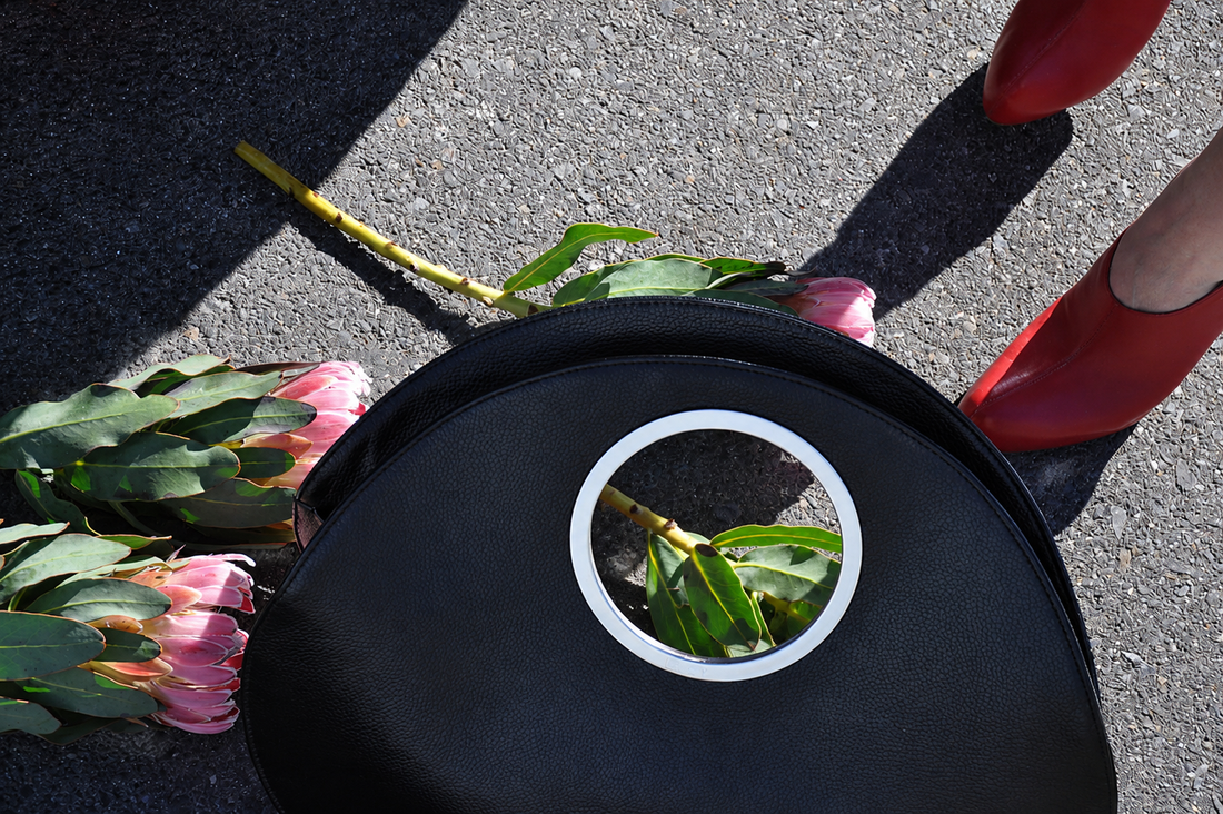 black LUCY bag laid flat on dark cobblestone, surrounded by pink protea flowers, red heeled shoe visible at the edge, overhead shot.