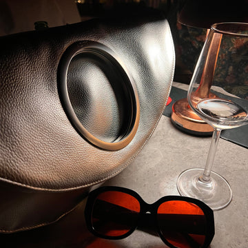 Evening lifestyle — Metallic Silver bag close-up on restaurant table with wine glass and sunglasses
10. Interior detail — cobalt blue lining, YKK zip and LUCY branded leather medallion keychain
11. Hardware close-up — branded LUCY silver ring detail on Metallic Black bag
12. Iridescent hands shot — close-up of hands holding Metallic Black bag, dark editorial setting