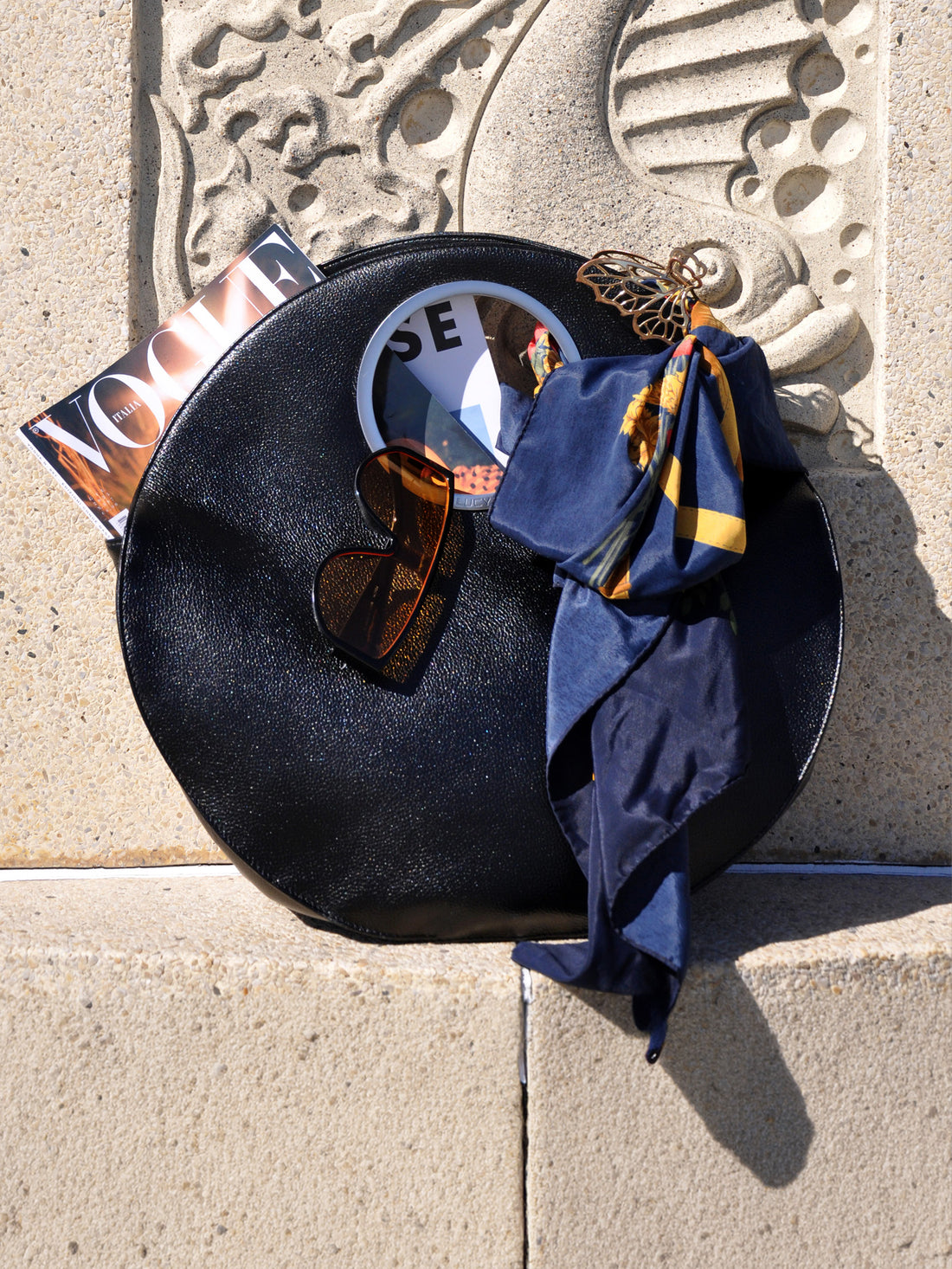 black Lucy bag against warm stone wall with Vogue magazine, silk scarf and sunglasses