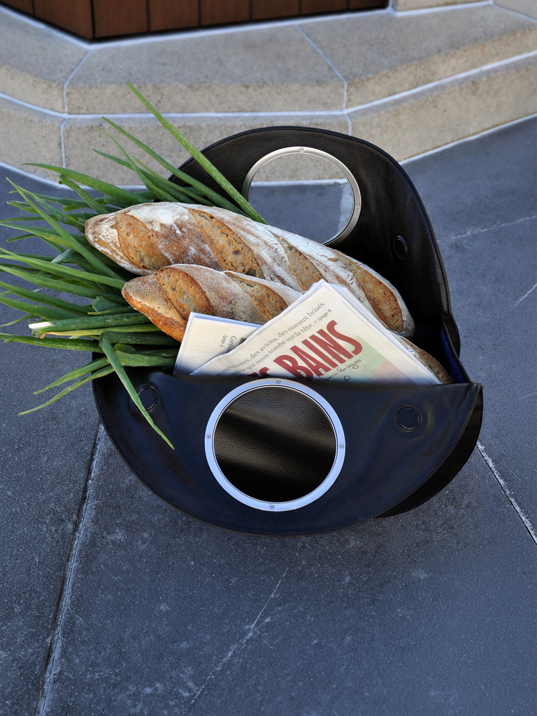 black Lucy bag as tote on grey slate tiles, filled with baguette, green onions and a newspaper