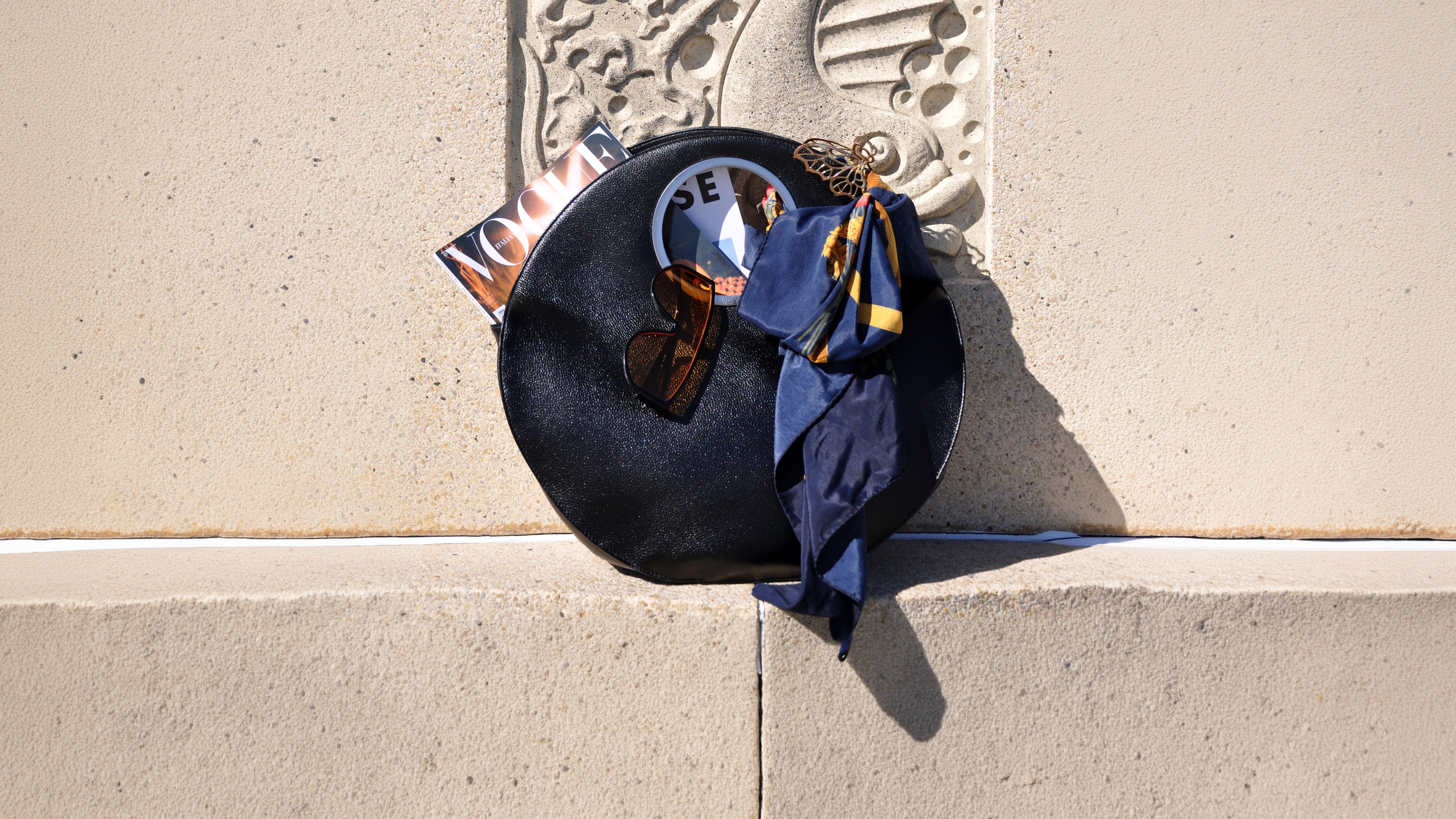 black Lucy bag against warm stone wall with Vogue magazine, silk scarf and sunglasses