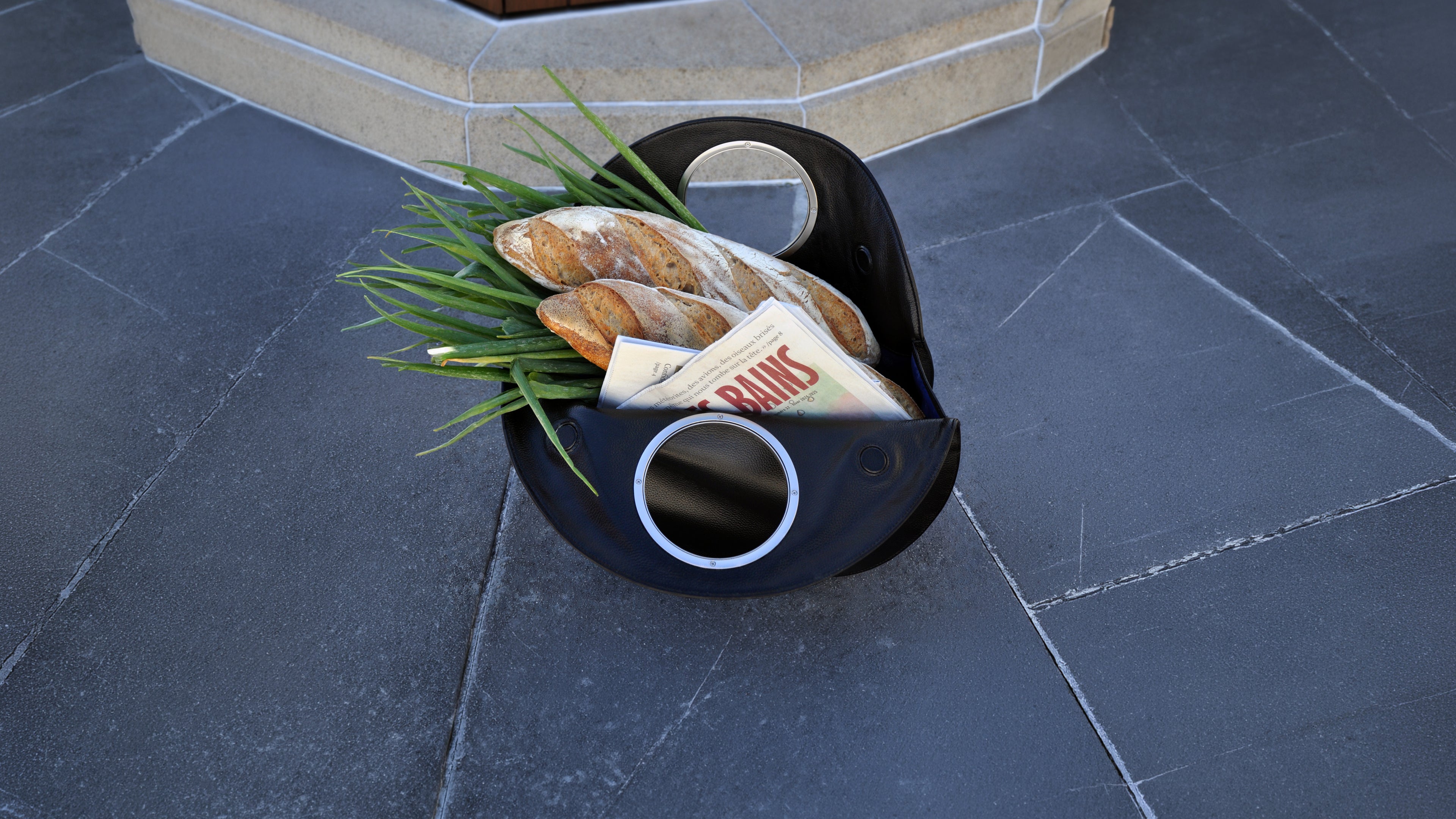 black Lucy bag as tote on grey slate tiles, filled with baguette, green onions and a newspaper